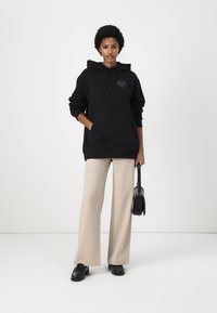 Black hooded sweatshirt with a front pocket and a tonal heart logo. Paired with beige wide-leg pants and black shoes, accessorized with a bag.