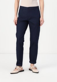 Pantalone od pamuka u tamnoplavoj boji s ravnim nogavicama, s kopčanjem na gumb, petljama za remen i zavrnutim manžetama. Uklopljeni s obućom svijetlih boja.