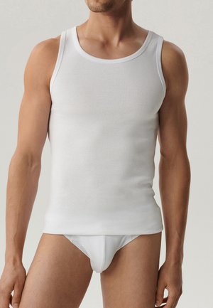 Mężczyzna w białej podkoszulce na ramiączkach i białych majtkach, stojący na tle jasnego jednolitego tła, tors widoczny od szyi do połowy uda.