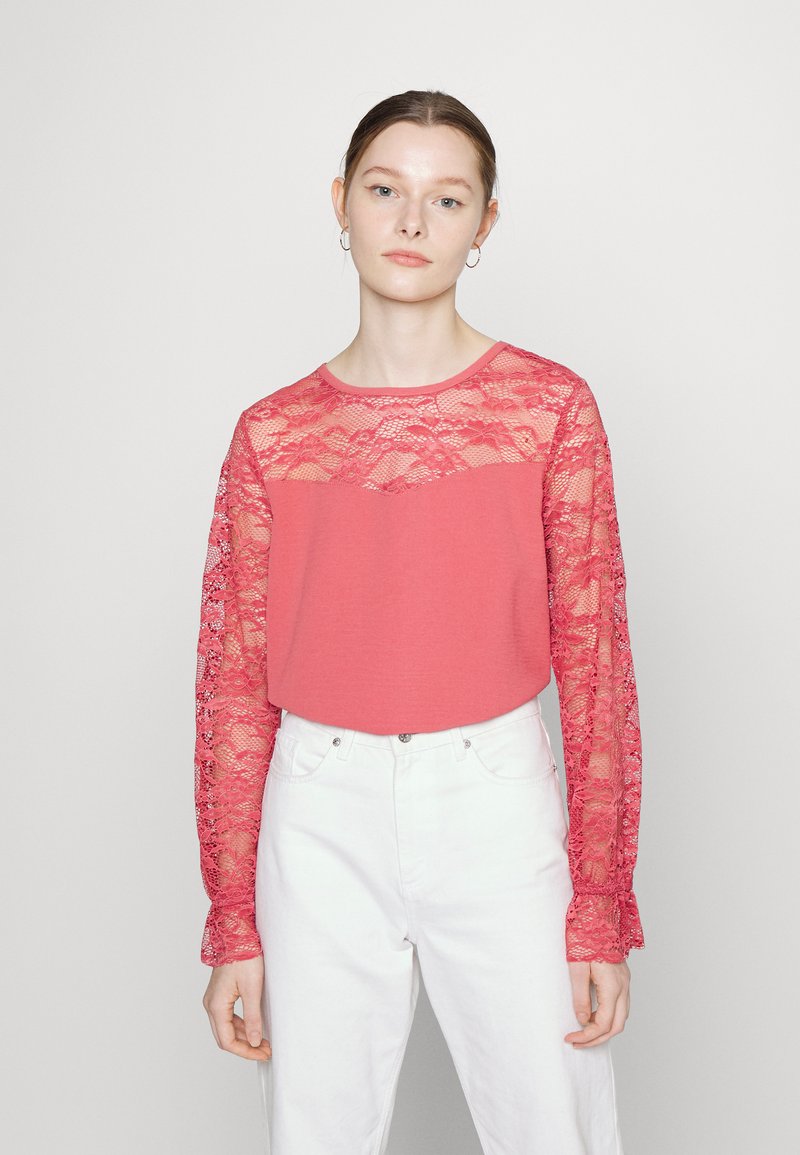 Blusa longa vermelha de mangas de renda e corpete, com decote redondo e textura suave. Combinada com calças brancas de cintura alta.