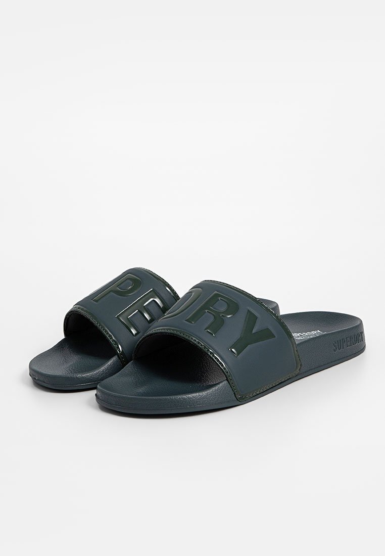 Sandales à glissière vert foncé avec une finition brillante, arborant une large sangle emboutie sur laquelle est inscrit le texte "SUPERDRY" en lettres majuscules contrastantes.