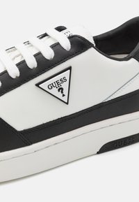 Sneaker bianco e nero realizzato in pelle sintetica con un logo triangolare. Presenta lacci bianchi, superficie testurizzata e suola in gomma.