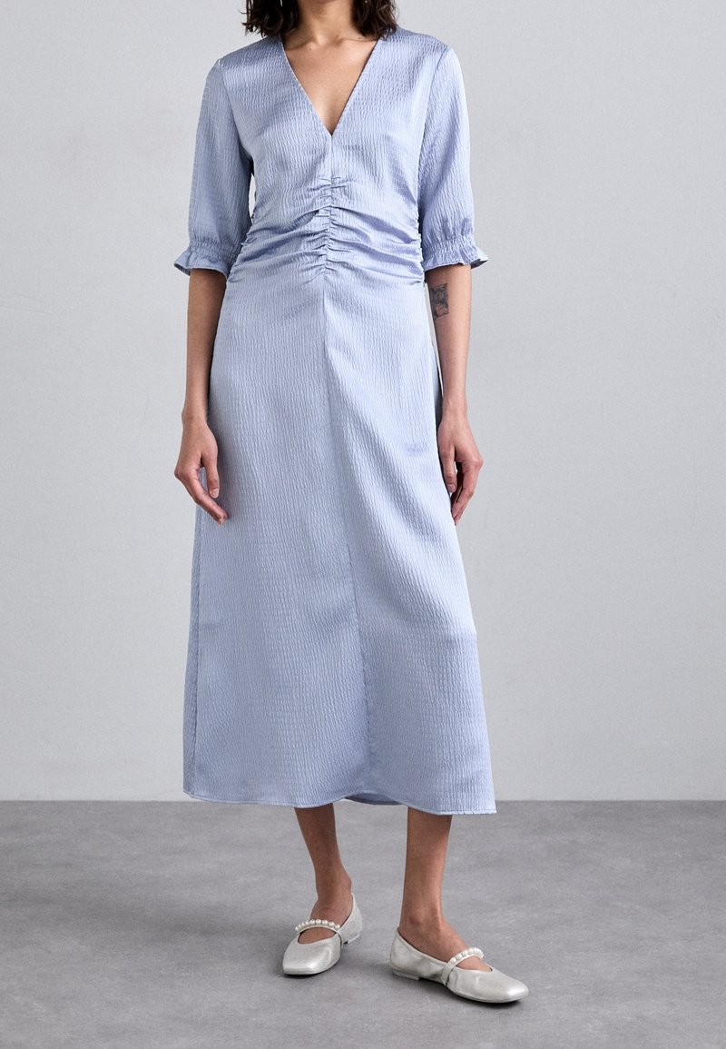 Femme portant une robe midi bleu clair froncée avec manches au coude et des chaussures plates argentées à brides perles, debout sur un sol gris avec un mur de fond gris.