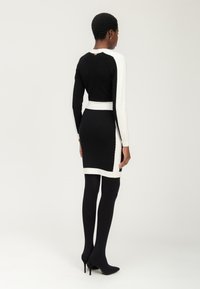 Strickkleid mit taillierter Silhouette, schwarzem Oberteil und kontrastierenden weißen Ärmeln. Verziert mit einem Taillenbund und spitzen Absätzen.
