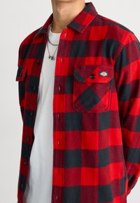 Chemise en flanelle à carreaux rouges et noirs avec une patte de boutonnage à l'avant, deux poches poitrine et un écusson de marque sur la poche gauche.