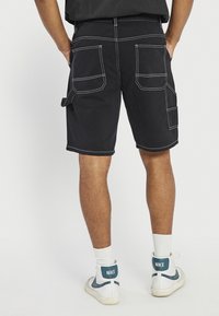 Mann trägt schwarze Shorts mit weißen Nähten und mehreren Taschen, steht mit den Händen in den hinteren Taschen, weiße Nike-Sneaker und weiße Socken.