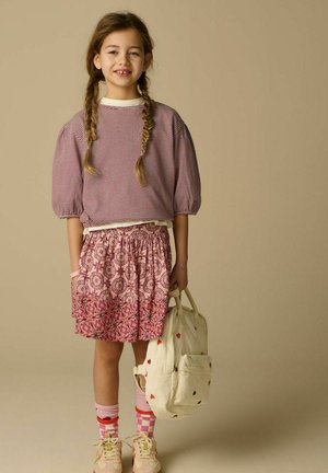 Jeune fille aux cheveux tressés portant un haut rayé et une jupe rose à motifs, tenant un sac à dos crème avec des motifs de cœurs.