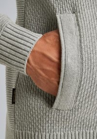 Grijze textuurtrui met geribde boorden en een zijzak, met een geweven patroon en een klein zwart label.