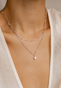 Collares en capas de plata con eslabones circulares y trenzados, con pequeños acentos de perlas y un colgante ovalado de perla, llevados sobre una blusa clara.