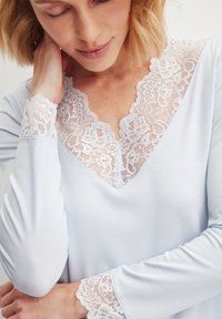 Lichtblauw langarmtop met een V-hals, voorzien van fijne witte kantaccenten bij de halslijn en manchetten. Zachte stoftextuur.