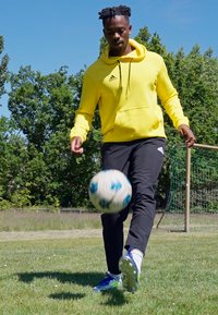 Gul hoodie med en frontlomme, sorte træningsbukser og blå fodboldstøvler. En fodbold med blå detaljer bevæger sig på græsset.
