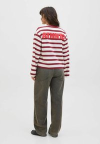 Model staat met de rug naar de camera, gekleed in een gestreepte lange mouwen shirt met "ATHLETIC" in vette rode letters op de achterkant, in combinatie met wijde donkere broek.