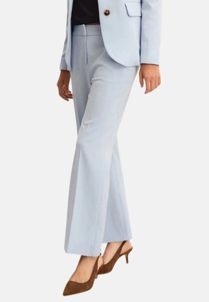 Donna che indossa pantaloni azzurri su misura e blazer abbinato, abbinati a décolleté marroni in camoscio con tacco basso, in piedi davanti a uno sfondo bianco.