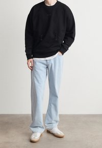 Persona che indossa una felpa nera, una maglietta bianca, jeans azzurri e scarpe da ginnastica bianche, in piedi su un pavimento lucido contro una parete bianca.