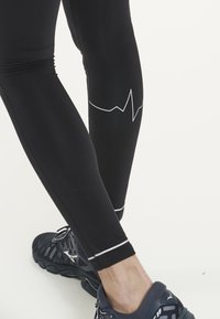 Sorte træningsleggings med en tætsiddende design, der har et hvidt hjerterytmemønster på læggen og en diskret hvid detalje ved anklen.