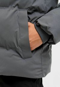 Giacca imbottita grigia con finitura liscia e lucida. Presenta una tasca laterale e dettagli a coste sul polsino. Vista ravvicinata che mostra il braccio e la mano chiusa.