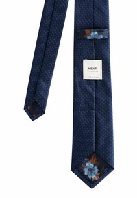 Tumši zils kaklasaites ar maziem oranžiem punktiem, ziedu auduma cilpa un gals, kā arī balta birka ar uzrakstu "NEXT Tailoring Made in China."