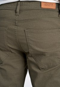 Pantalons vert olive en tissu texturé, comportant des poches arrière et une étiquette en cuir portant la mention "JACK'S SPORTSWEAR INTL."