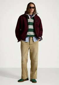 Homme portant une veste bordeaux, un polo de rugby rayé vert et blanc, un pantalon kaki, des baskets vertes et des lunettes de soleil, se tenant avec les mains dans les poches.