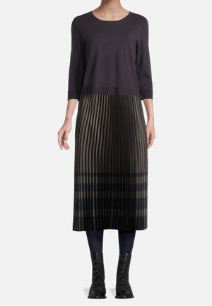 Žena oblečená v černém tričku s rukávy po lokty, plisované midi sukně s vodorovnými pruhy, tmavé legíny a černé kozačky s tkaničkami.