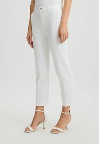 Pantalones de mezclilla blancos con un diseño ajustado, que cuentan con un cierre de botón, dos bolsillos laterales y un corte corto. Combinados con sandalias de tacón transparente.