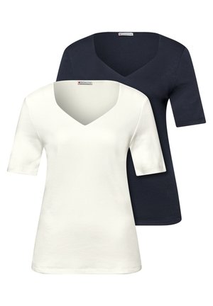 Dwie kobiety stojące, ubrane w codzienne dżinsy i kurtki; jedna trzyma beżową torbę na ramię, obie uśmiechają się na tle gładkiego beżowego tła.