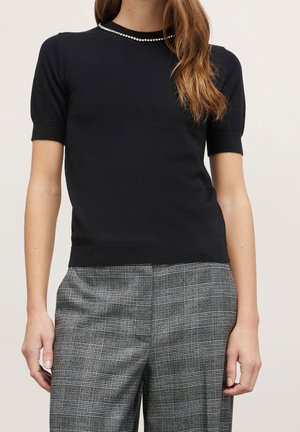 Kvinde iført kortærmet sort sweater med rhinsten ved halsudskæringen og højtaljede grå ternede bukser, stående mod en ensartet baggrund.