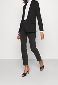 Mustat bleiserit, joissa on viistot kaulukset, yhdistettynä slim fit -tummanharmaisiin farkkuihin ja mustiin korkokenkiin. Valkoinen paita bleiserin alla.