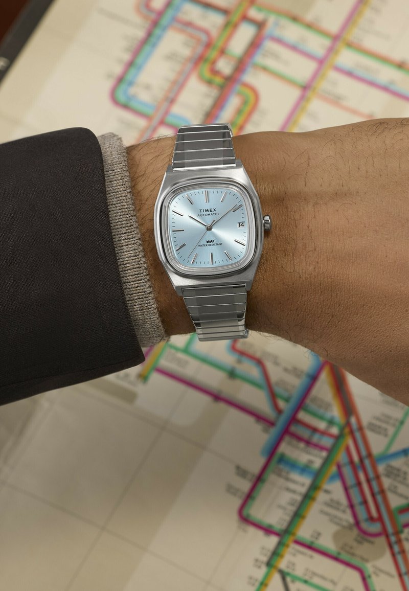 Καρπό με ασημένιο αυτόματο ρολόι Timex με μπλε καντράν που δείχνει 10:13, φόντο πολύχρωμου χάρτη συγκοινωνιών με διάφορες διαδρομές και στάσεις.