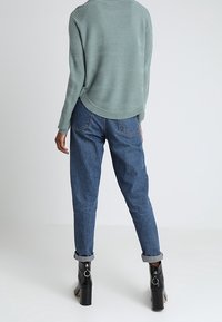 Pull vert clair texturé avec un ourlet incurvé, associé à un jean bleu taille haute et des bottines à talons chunky noires, avec les poignets retroussés.