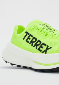 Neon zöld futócipő fekete "TERREX" felirattal, texturált szintetikus felsőrésszel, vastag fehér középtalppal és durva fekete talppal a tapadás érdekében.