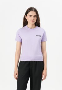 Vijolična bombažna majica s kratkimi rokavi, okroglim izrezom, ravnimi šivi in majhnim črnim logotipom na levi strani prsi. Kombinirana s črnimi hlačami.