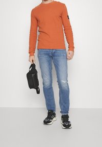 Oranžová košile s dlouhým rukávem z texturované látky, světle modré děrované džíny a černé tenisky, držící malou černou kabelku.