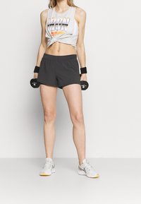 Camisa sin mangas gris claro con gráfico de "IRON", pantalones cortos gris oscuro, muñequeras negras y zapatillas deportivas blancas con suelas amarillas.