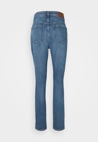 Zadní pohled na modré džíny s rovnými nohavicemi, které mají dvě nášivkové kapsy a hnědou koženou značkovou nášivku na pásku.