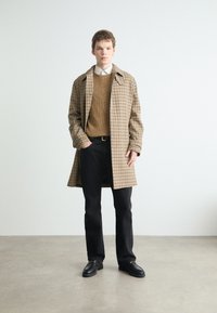 Braun karierten Mantel mit Kragen über einem strukturierten braunen Pullover. Schwarze Hosen und schwarze Schuhe runden das Outfit ab. Einfaches, minimalistisches Design.