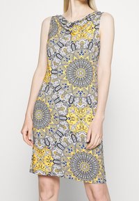 Robe sans manches avec un motif orné jaune, noir et bleu comprenant des dessins floraux et géométriques. Tissu lisse avec un col bénitier.