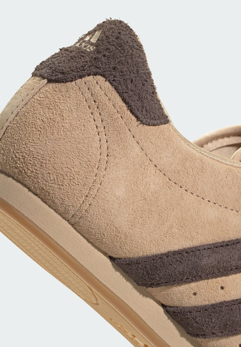 Zapatilla de ante beige con acentos en marrón oscuro y tres franjas distintivas. Presenta una parte superior texturizada y suela de goma lisa. Logo dorado en el talón.