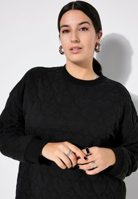 Zwart gewatteerd sweat-shirt met hartvormige patronen, ronde halslijn en geribde manchetten. Model toont ringen op de handen tegen een neutrale achtergrond.