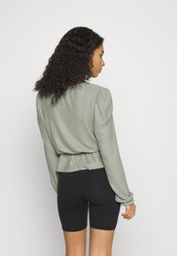 Blusa de mangas longas, verde claro, com padrão texturizado, cintura-elástica e mangas franzidas, combinada com calções pretos.