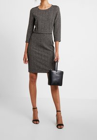 Grå mønstret kjole med trekvart ærmer og en tætsiddende silhuet. Sort håndtaske med bærehank. Sorte åben tå hæle.