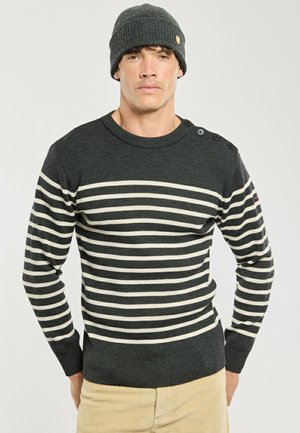 Uomo che indossa un maglione a righe grigio scuro e bianco con bottoni sulla spalla e un berretto di lana grigio scuro.