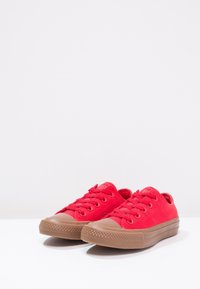 Zapatillas de lona rojas con punteras redondeadas, cordones planos, suelas de goma y acentos marrones texturizados en la puntera y en el borde de la suela.