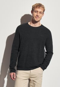 Pull long noir texturé à manches longues avec un col arrondi. Associé à un pantalon beige clair. Présente un logo de marque subtil sur la poitrine.
