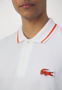 Polo shirt blanche avec un tissu texturé, un col classique avec des rayures orange, et un petit logo de crocodile rouge brodé sur la poitrine.