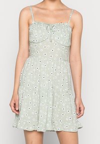 Krátké zelené květinové šaty s tenkými ramínky, s nařaseným živůtkem, volánkovým lemem a malými bílými květinami s černými středy.