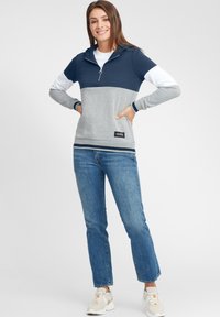 Felpa grigia e blu con zip, maniche lunghe, dettagli bianchi e patch con logo. Indossata con jeans blu e sneakers chiare.