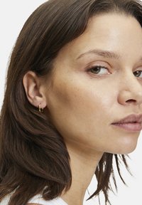 Orecchini a cerchio d'oro adornano l'orecchio di un modello. Gli orecchini sono piccoli, circolari e lucenti, che si abbinano ai capelli castano medio e a un tono di pelle neutro.