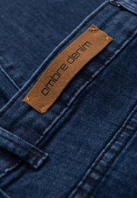 Tissu denim bleu foncé avec une étiquette en cuir marron cousue à l'arrière, affichant le texte "ombre denim" en noir.