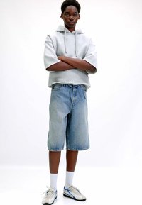Haut de survêtement gris, shorts en denim bleu et baskets grises avec accents bleus. Le mannequin se tient les bras croisés sur un fond blanc.